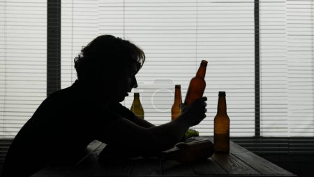 Photo for In the frame a young man sits at a table in a cafe. Nearby are empty bottles of alcohol. He is looking at one of them and thinking about something. Demonstrates loneliness, alcoholism, sadness. - Royalty Free Image