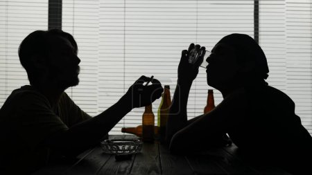 Foto de Jóvenes sentados en una mesa en un establecimiento. Rodeado de botellas vacías. Uno enciende un cigarrillo. El otro termina su bebida de su vaso. Están hablando de algo. Tiro medio. - Imagen libre de derechos