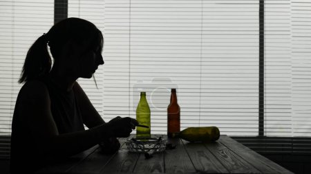 Photo for In the frame, a young woman sits on a table surrounded by empty bottles. She demonstrates alcoholic intoxication. She smokes a cigarette, after finishing it she puts it out. Shes lonely, sad. - Royalty Free Image