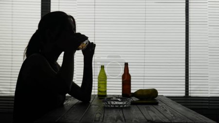 Photo for In the frame of a young woman she sits in a cafe at a table against a background of empty bottles of alcohol. She sits alone and drinks alcohol from a glass, thinking about something. She is sad. - Royalty Free Image