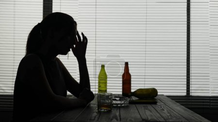 Photo for In a silhouette of a young woman sitting in a cafe at a table on the background of empty bottles of alcohol and a pack of cigarettes. She is sitting alone, she is upset, she is sad. Medium shot. - Royalty Free Image