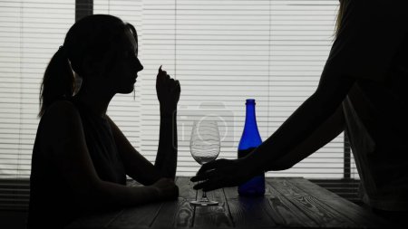 Photo for In the frame in silhouette of a young woman she is sitting in a cafe at a table. The waiter has brought her alcohol and a glass. She sits alone and is going to drink. Demonstrates alcoholism, sadness. - Royalty Free Image