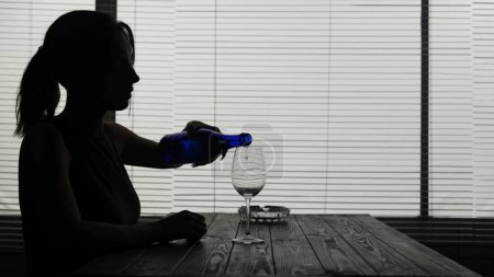 Photo for In the frame in silhouette of a young woman she is sitting in a cafe at a table. She pours herself alcohol into a glass and is about to drink alone. Demonstrates alcoholism, sadness. Medium shot. - Royalty Free Image