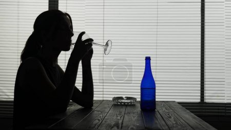 Photo for In the silhouette of a young woman sitting at a table in a cafe thinking about something. She is drinking alcohol, possibly wine from a glass alone. Demonstrates alcoholism, sadness. Medium shot. - Royalty Free Image