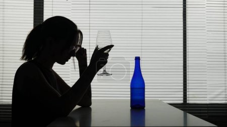 Photo for A young woman with glasses is sitting at a table in a cafe thinking about something, a bottle and an ashtray are next to her. She is holding a glass of alcohol. Demonstrates loneliness, alcoholism. - Royalty Free Image