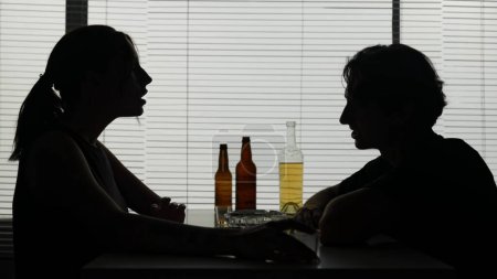 Téléchargez les photos : Un homme et une femme sont à table dans les bouteilles environnantes. Sur une table les verres à vin coûtent avec l'alcool. Démontrez-lui qu'il boit de l'alcool. Une femme allume une cigarette et lui dit :. - en image libre de droit
