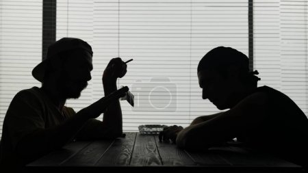 Téléchargez les photos : La vidéo montre deux jeunes hommes assis à une table dans un café. L'un fume et tient un sac de drogue. Ils se sont rencontrés pour une transaction. Ils parlent de quelque chose et regardent le sac. - en image libre de droit