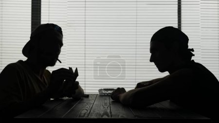 Photo for Two young men are sitting at a table in a cafe. One is smoking and holding a wad of money looks at them and counts. Theyve met for a transaction. They talk about something and look at the bag. - Royalty Free Image