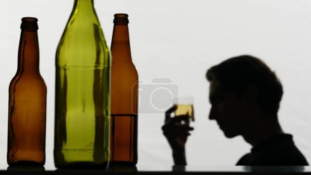 Photo for In the close-up shot, there are empty, broken glass bottles full of alcohol. In the background, a strong man holds a glass of alcohol in his hands and examines it. Then he drinks sad and sad. - Royalty Free Image