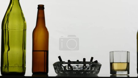 Foto de Un primer plano de botellas de vidrio vacías con alcohol, un cenicero y un vaso lleno de alcohol sobre un fondo blanco. Demuestra malos hábitos, adicción. Tiro medio. - Imagen libre de derechos
