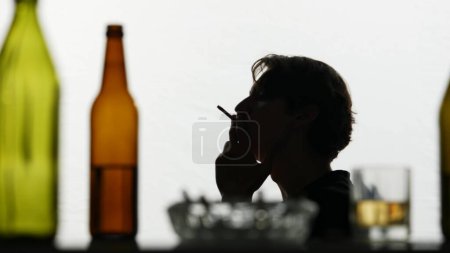 Téléchargez les photos : Dans le gros plan, la caméra montre des bouteilles en verre vides, dissipées, avec de l'alcool, des cendres avec des cigarettes et un verre rempli d'alcool. En arrière-plan, un homme allume une cigarette et fume. - en image libre de droit