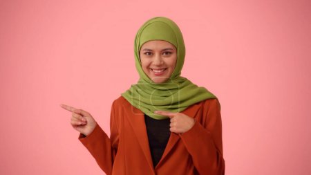Téléchargez les photos : Photo isolée de taille moyenne capturant une jolie jeune femme portant un hijab, voile. Elle montre du doigt le côté gauche du cadre. Place pour votre publicité, maquette, promotionnel. - en image libre de droit