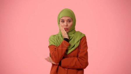 Foto de Foto aislada de tamaño mediano que captura a una atractiva joven con un hiyab, velo. Ella expresa sorpresa y conmoción, tocándose la barbilla. Lugar para su anuncio, cultural, diversidad. - Imagen libre de derechos