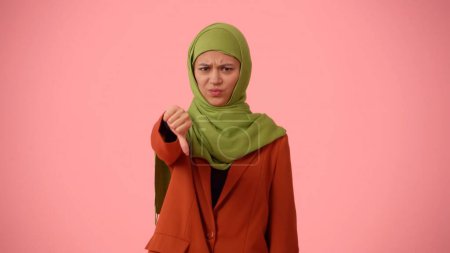 Foto de Foto aislada de tamaño mediano que captura a una atractiva joven con un hiyab, velo. Señala con el pulgar hacia abajo en desacuerdo. Lugar para su anuncio, cultural, diversidad. - Imagen libre de derechos