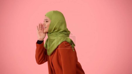 Téléchargez les photos : Photo isolée de profil latéral moyen capturant une jolie jeune femme portant un hijab, voile. Elle a l'air sournoise, couvrant sa bouche, racontant un secret. Place pour votre publicité, culturel, diversité. - en image libre de droit
