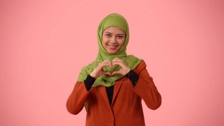 Téléchargez les photos : Photo isolée de taille moyenne capturant une jolie jeune femme portant un hijab, voile. Elle fait un cœur avec ses mains et sourit. Place pour votre publicité, diversité culturelle. - en image libre de droit