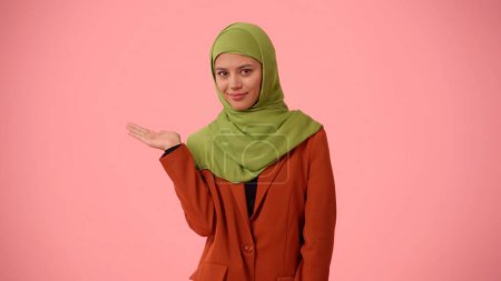 Téléchargez les photos : Photo isolée de taille moyenne capturant une jolie jeune femme portant un hijab, voile. Elle pointe sa main vers le côté gauche du cadre. Place pour votre publicité, maquette, promotionnel. - en image libre de droit