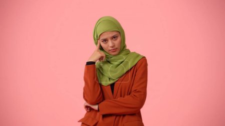 Foto de Foto aislada de tamaño mediano que captura a una atractiva joven con un hiyab, velo. Ella inclina su cabeza sobre su dedo, confundida y cansada. Lugar para su anuncio, cultural, diversidad. - Imagen libre de derechos