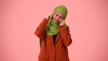 Téléchargez les photos : Photo isolée de taille moyenne capturant une jolie jeune femme portant un hijab, voile. Elle se colle les oreilles avec les doigts en désaccord. Place pour votre publicité, culturel, diversité. - en image libre de droit