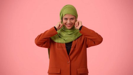 Photo for Medium-sized isolated photo capturing an attractive young woman wearing a hijab, veil. She is plugging her ears with fingers and smiling widely. Place for your advertisement, cultural, diversity. - Royalty Free Image