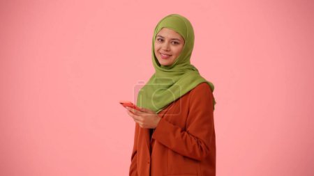 Téléchargez les photos : Photo isolée de taille moyenne capturant une jolie jeune femme portant un hijab, voile. Elle tient un smartphone et sourit à la caméra. Place pour votre publicité, culturel, diversité. - en image libre de droit