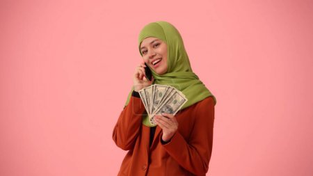 Foto de Foto aislada de tamaño mediano que captura a una atractiva joven con un hiyab, velo. Ella está hablando por teléfono y mostrando billetes de dólar en la cámara. Lugar para publicidad, cultura, diversidad. - Imagen libre de derechos