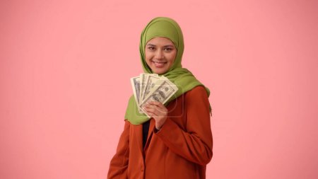Photo for Medium-sized isolated photo capturing an attractive young woman wearing a hijab, veil. She is taking photos of herself with dollar bills in her hand. Place for advertisement, diversity, influencer. - Royalty Free Image