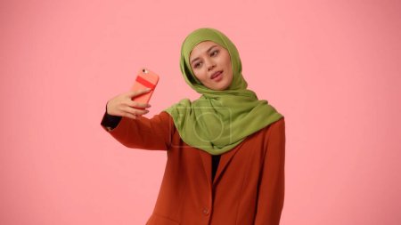 Téléchargez les photos : Photo isolée de taille moyenne capturant une jolie jeune femme portant un hijab, voile. Elle prend des selfies, des photos d'elle-même avec le smartphone. Lieu de publicité, de culture, de diversité. - en image libre de droit