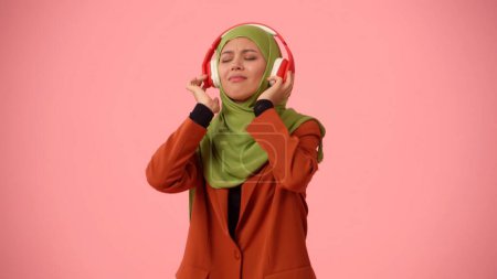 Foto de Foto aislada de tamaño mediano que captura a una atractiva joven con un hiyab, velo. Está escuchando música enérgica a través de auriculares y bailando. Lugar para publicidad, cultura, diversidad. - Imagen libre de derechos