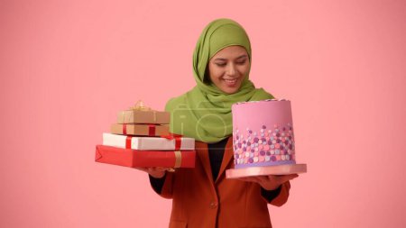 Foto de Foto aislada de tamaño mediano que captura a una atractiva joven con un hiyab, velo. Ella está sosteniendo regalos y un pastel en las manos, emocionada y feliz. Lugar para su anuncio, vacaciones, diversidad. - Imagen libre de derechos