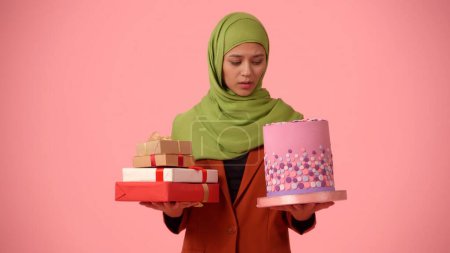 Photo for Medium-sized isolated photo capturing an attractive young woman wearing a hijab, veil. She is holding gifts and a cake in her hands, frustrated. Place for your advertisement, holidays, diversity. - Royalty Free Image