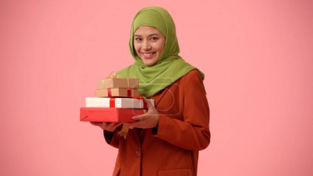 Téléchargez les photos : Photo isolée de taille moyenne capturant une jolie jeune femme portant un hijab, voile. Elle tient des boîtes-cadeaux dans ses mains, excitée et heureuse. Lieu pour votre publicité, vacances, diversité. - en image libre de droit