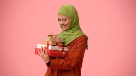 Téléchargez les photos : Photo isolée de taille moyenne capturant une jolie jeune femme portant un hijab, voile. Elle tient des boîtes-cadeaux dans ses mains, excitée et heureuse. Lieu pour votre publicité, vacances, diversité. - en image libre de droit