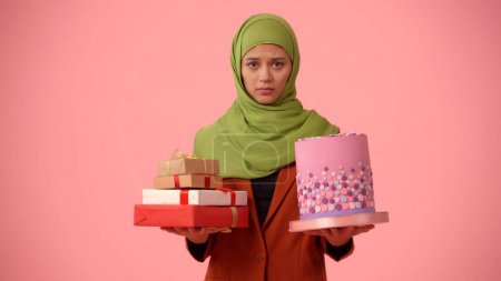 Foto de Foto aislada de tamaño mediano que captura a una atractiva joven con un hiyab, velo. Ella sostiene regalos y un pastel en sus manos, frustrada. Lugar para su anuncio, vacaciones, diversidad. - Imagen libre de derechos