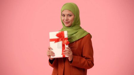 Foto de Foto aislada de tamaño mediano que captura a una atractiva joven con un hiyab, velo. Ella sostiene una caja de regalo en sus manos, emocionada y feliz. Lugar para su anuncio, vacaciones, diversidad. - Imagen libre de derechos