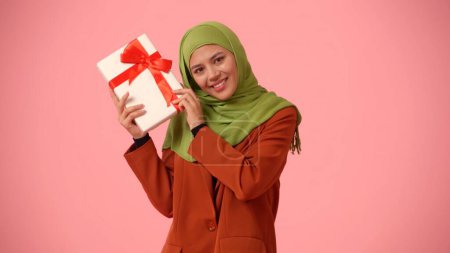 Photo for Medium-sized isolated photo capturing an attractive young woman wearing a hijab, veil. She is shaking a gift box in her hands, excited and happy. Place for your advertisement, holidays, diversity. - Royalty Free Image