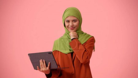 Foto de Foto aislada de tamaño mediano que captura a una atractiva joven con un hiyab, velo. Ella sostiene una tableta, un portátil, sonriendo a la cámara. Diversidad cultural, empleo y educación, publicidad. - Imagen libre de derechos