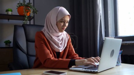 Foto de Foto de tamaño mediano que captura a una atractiva joven con un hiyab, velo. Ella está trabajando en una computadora portátil, escribiendo agresivamente como si estuviera loca. Diversidad cultural, empleo y educación, publicidad. - Imagen libre de derechos