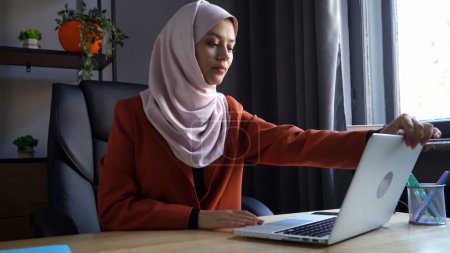 Foto de Foto de tamaño mediano que captura a una atractiva joven con un hiyab, velo. Abre su portátil para empezar a trabajar o estudiar. Parece contenta. Diversidad cultural, empleo y educación, publicidad - Imagen libre de derechos