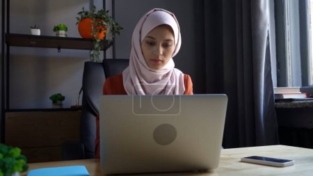 Foto de Foto de tamaño mediano que captura a una atractiva joven con un hiyab, velo. Ella está trabajando o estudiando en su computadora portátil, relajada y pacífica. Diversidad cultural, empleo y educación, publicidad. - Imagen libre de derechos