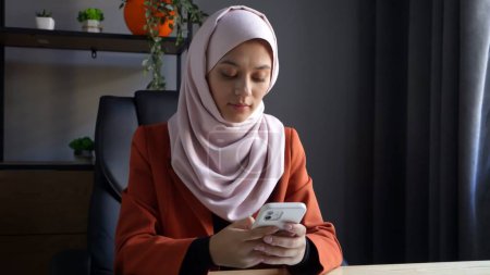Photo for Medium-sized photo capturing an attractive young woman wearing a hijab, veil. She is holding a smartphone, typing, searching for something online. Place for your advertisement, cultural, diversity. - Royalty Free Image
