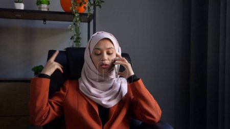 Foto de Foto de tamaño mediano que captura a una atractiva joven con un hijab, velo sentado en la silla de la oficina. Está teniendo una llamada de negocios. Diversidad cultural, empleo y educación, publicidad. - Imagen libre de derechos