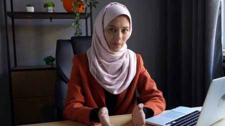 Téléchargez les photos : Photo de taille moyenne capturant une jolie jeune femme dans un hijab, voile assis devant l'ordinateur portable dans le bureau, parlant à la caméra. Diversité culturelle, emploi et éducation, publicité. - en image libre de droit