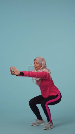 Téléchargez les photos : Photo isolée pleine grandeur capturant une jolie jeune femme portant un vêtement de sport et un hijab, Sheila. Elle fait de l'exercice, des squats. Place pour votre publicité, maquette, promotion, sport. - en image libre de droit