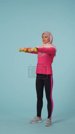Photo for Full-sized isolated photo capturing an attractive young woman wearing a sportswear and a hijab, sheila. She is exercising, lifting dumbbells. Place for your advertisement, mock up, promotional, sport. - Royalty Free Image