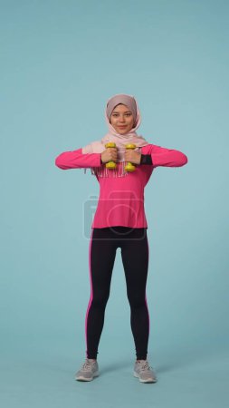 Foto de Foto aislada de tamaño completo que captura a una atractiva joven con ropa deportiva y un hiyab, Sheila. Ella está haciendo ejercicio, levantando pesas. Lugar para tu anuncio, maqueta, promoción, deporte. - Imagen libre de derechos