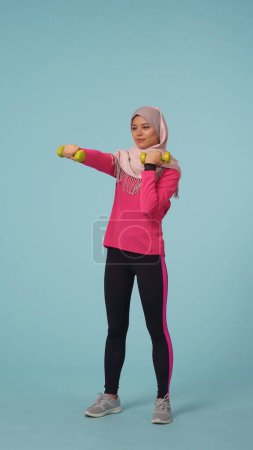 Foto de Foto aislada de tamaño completo que captura a una atractiva joven con ropa deportiva y un hiyab, Sheila. Ella está haciendo ejercicio con pesas, puñetazos. Lugar para tu anuncio, promoción, deporte. - Imagen libre de derechos