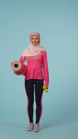 Foto de Foto aislada de tamaño completo que captura a una joven con ropa deportiva y un hiyab, Sheila. Ella sostiene pesas y hace ejercicio de alfombra, sonriendo. Lugar para tu anuncio, promoción, deporte. - Imagen libre de derechos