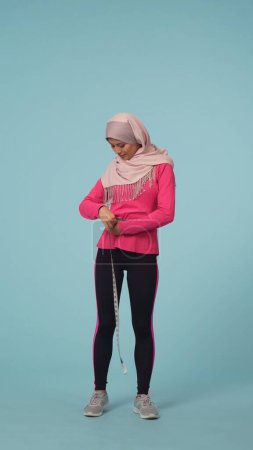 Foto de Foto aislada de tamaño completo que captura a una atractiva joven con ropa deportiva y un hiyab, Sheila. Está midiendo su cintura. Lugar para la publicidad, promocional, perder peso. - Imagen libre de derechos