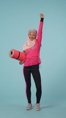 Foto de Foto aislada de tamaño completo que captura a una joven con ropa deportiva y un hiyab, Sheila. Ella sostiene una esterilla de ejercicio y salta en la emoción. Lugar para tu anuncio, promoción, deporte. - Imagen libre de derechos
